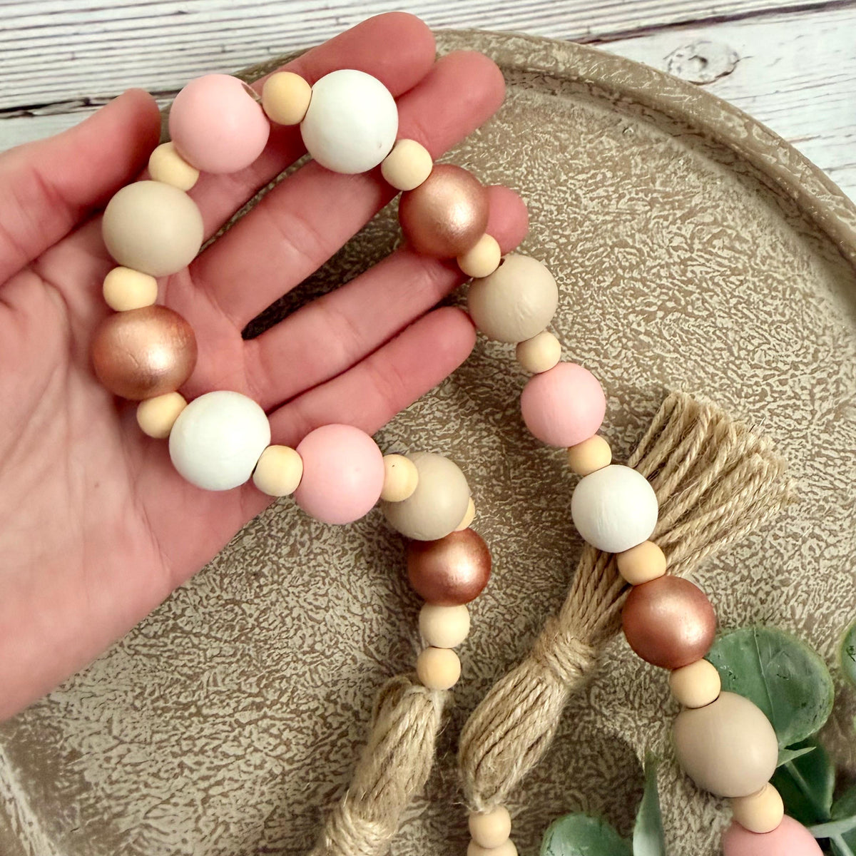 A wooden bead garland with pastel pink, white, beige, and metallic rose gold beads, held in a hand over a textured beige tray.