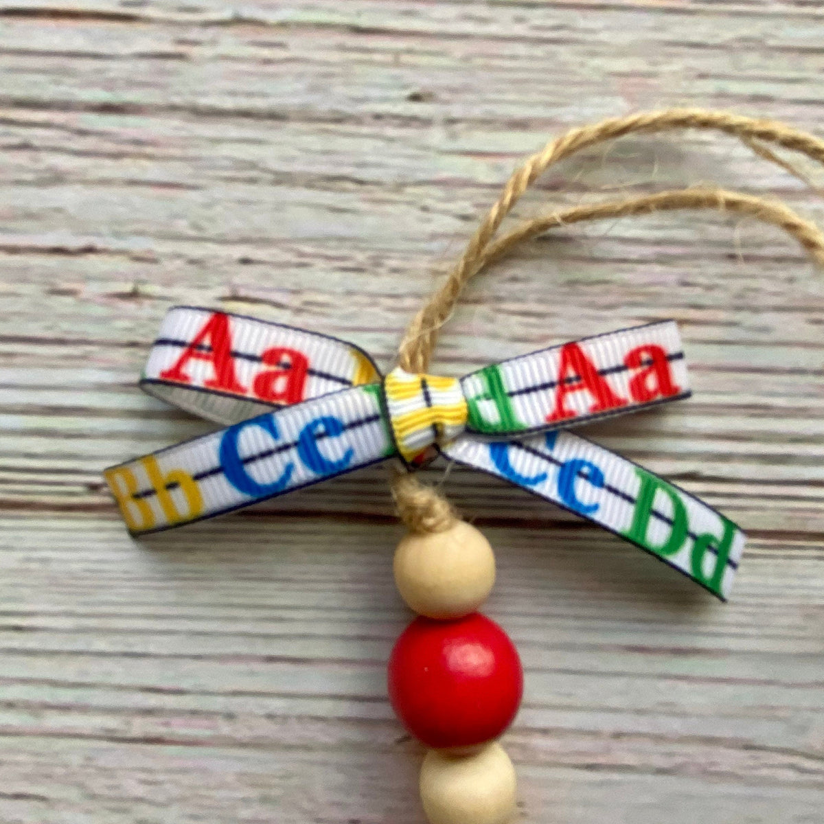 A handmade alphabet ornament featuring a bow made of printed alphabet ribbons spelling 'Aa Cc Dd Ee Ff Gg Hh Ii Jj Kk Ll Mm Nn Oo Pp Qq Rr Ss Tt Uu Vv Ww Xx Yy Zz', with three wooden beads (one red, two natural wood) and a jute hanging string, displayed on a light gray wood background.