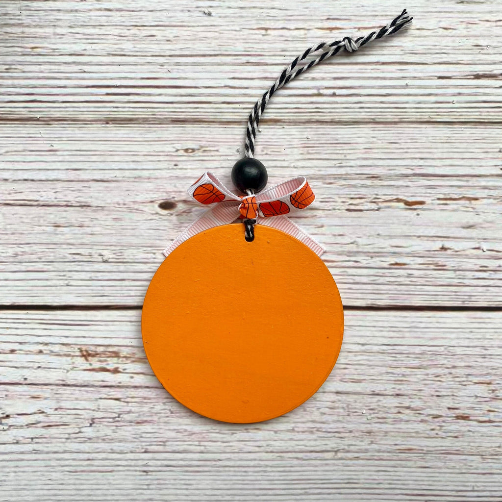An orange circular pumpkin-shaped ornament with a black and white twisted string hanger, a black bead, and a small orange and white patterned ribbon bow, displayed on a rustic white wood plank background.