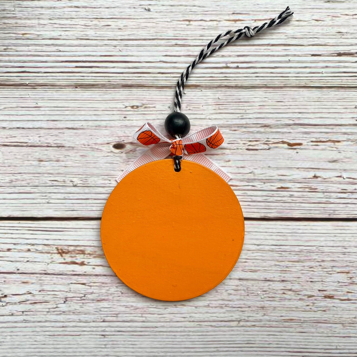 An orange circular pumpkin-shaped ornament with a black and white twisted string hanger, a black bead, and a small orange and white patterned ribbon bow, displayed on a rustic white wood plank background.