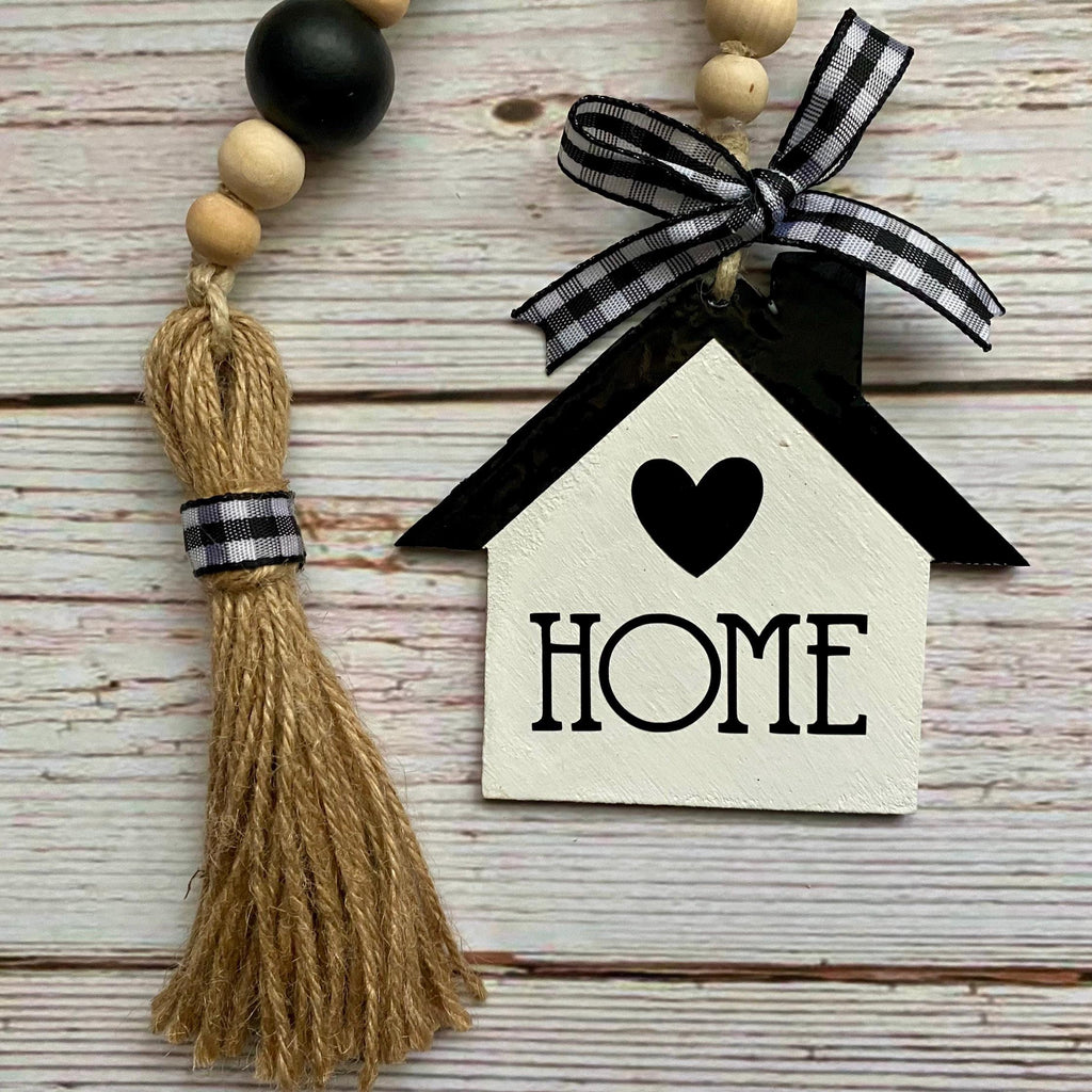 A rustic wooden bead garland with a tassel and a house-shaped ornament that says HOME, displayed on a whitewashed wood background.