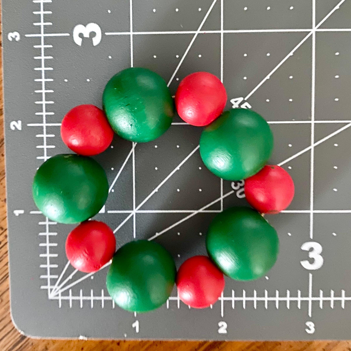 A small beaded wreath made of alternating glossy green and red round beads, placed on a gray cutting mat with measurement markings.