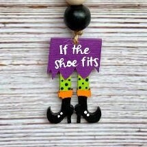 A Halloween-themed hanging ornament with a purple sign reading 'If the shoe fits' and legs in green and orange socks with black shoes, shown against a whitewashed wood background.