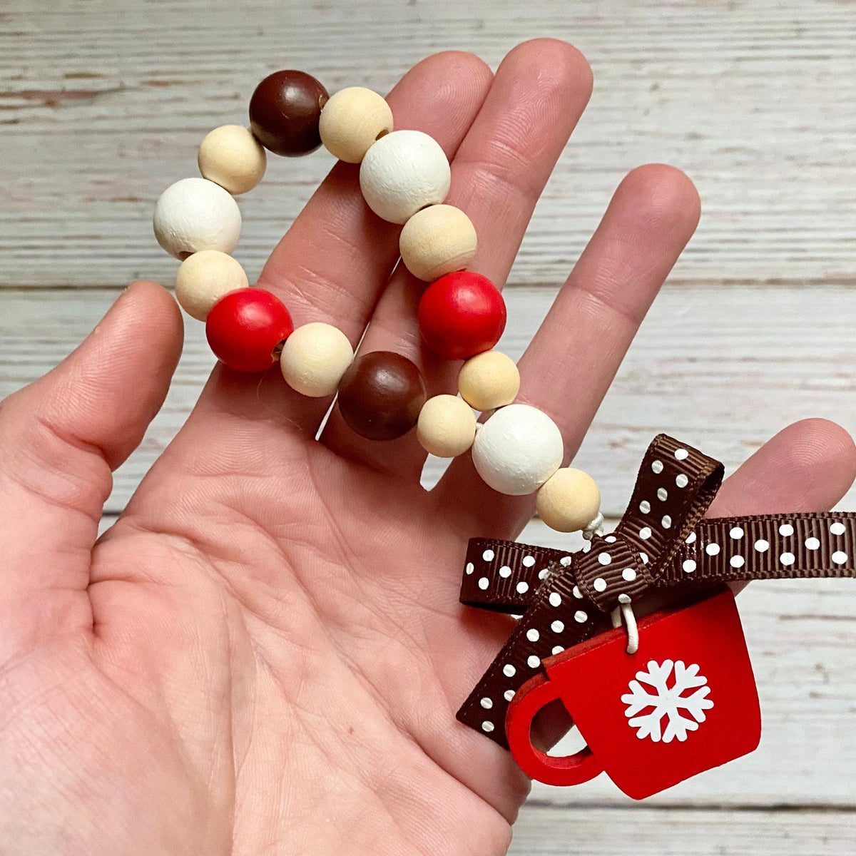 A hand holding a beaded holiday garland with red, brown, cream, and white wooden beads, featuring a brown polka dot ribbon bow and a red mug charm with a white snowflake, set against a light wood plank background.