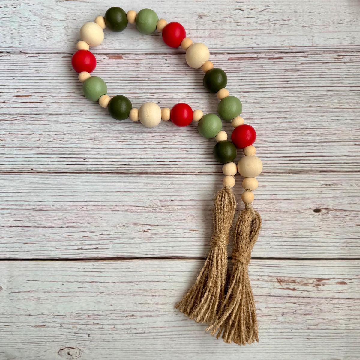 A beaded garland with alternating red, green, and natural wood beads, finished with two knotted jute tassels, laid flat on a whitewashed wooden surface.