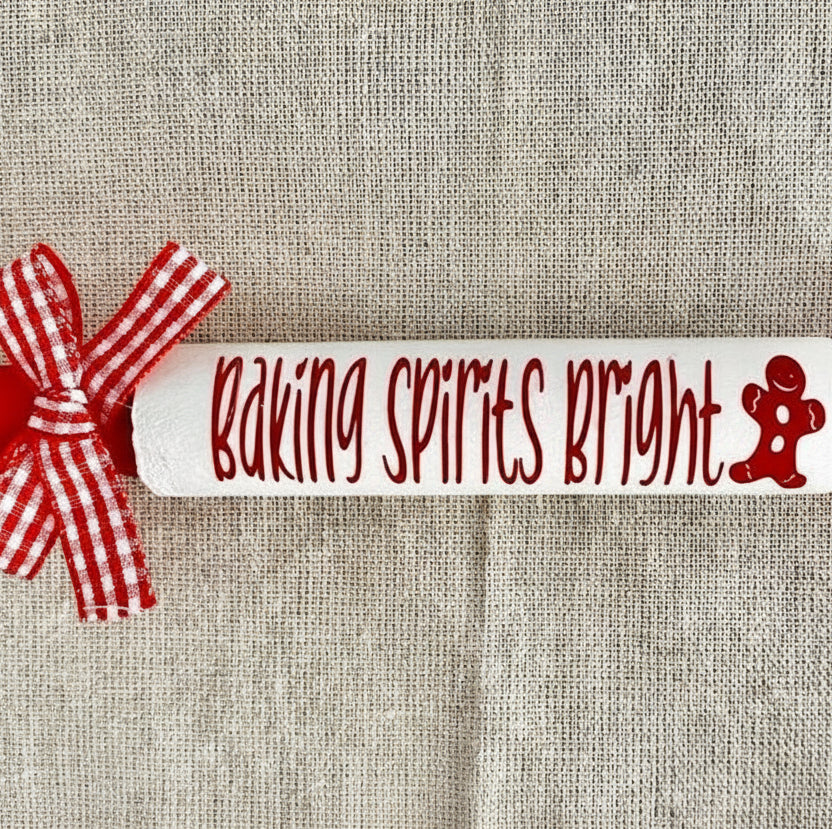 A white rolling pin with red handles and a red-and-white gingham ribbon bow, placed on a beige linen background. The center of the rolling pin reads 'Baking Spirits Bright' in red script with a small red gingerbread figure icon.