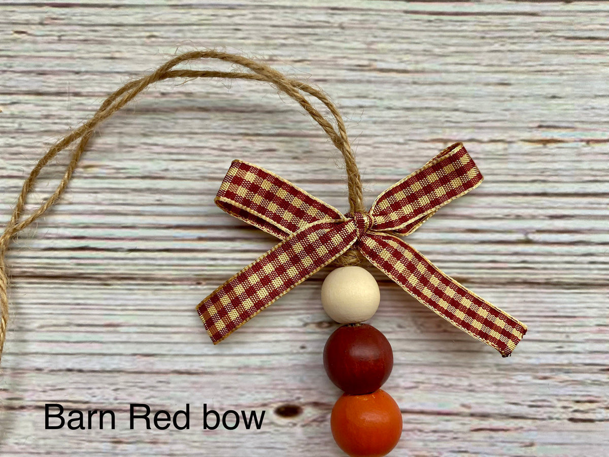 A rustic Christmas ornament featuring a barn red and tan gingham ribbon bow with three wooden beads below it, displayed on a whitewashed wood background. Text overlay reads 'Barn Red bow'.
