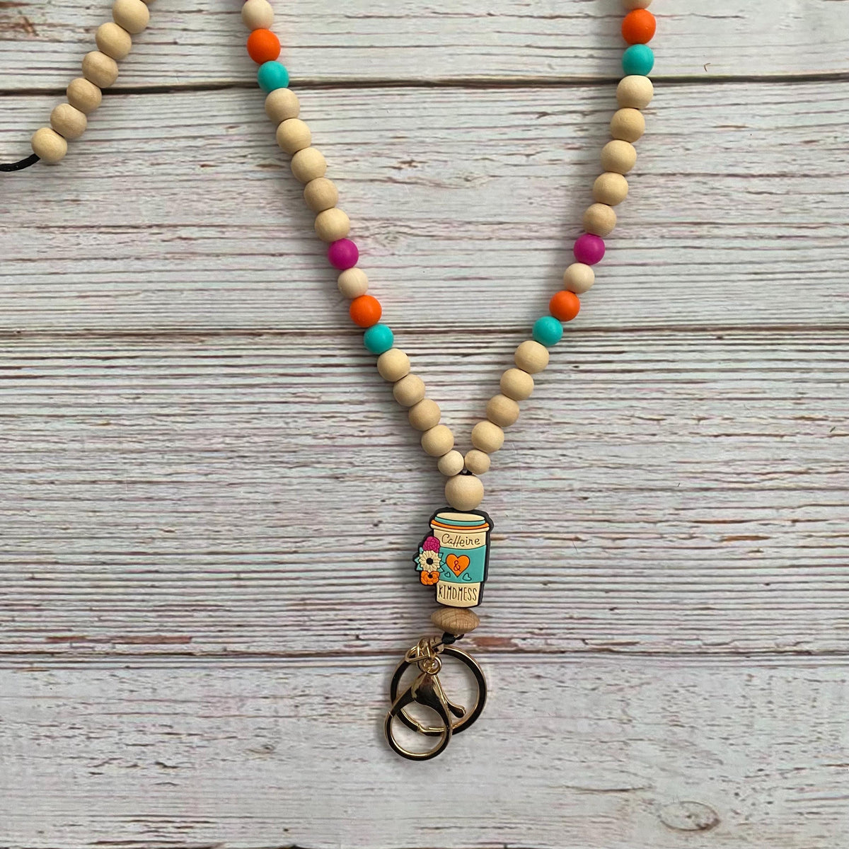 A beaded necklace with natural wood beads, colorful accent beads, and a central charm shaped like a takeout coffee cup. The necklace is displayed on a rustic, whitewashed wooden surface.