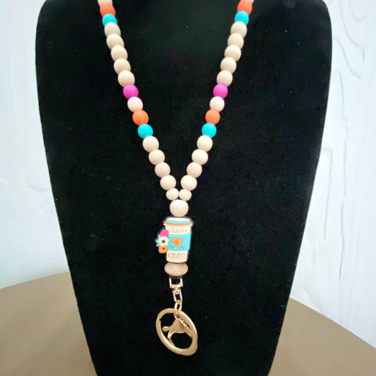 A beaded necklace displayed on a black velvet jewelry bust. The necklace features light beige wooden beads, alternating rows of small colorful beads in orange, turquoise, pink, and purple, a central silicone charm depicting a coffee cup, and a gold-tone metal clasp with a dog-shaped charm.