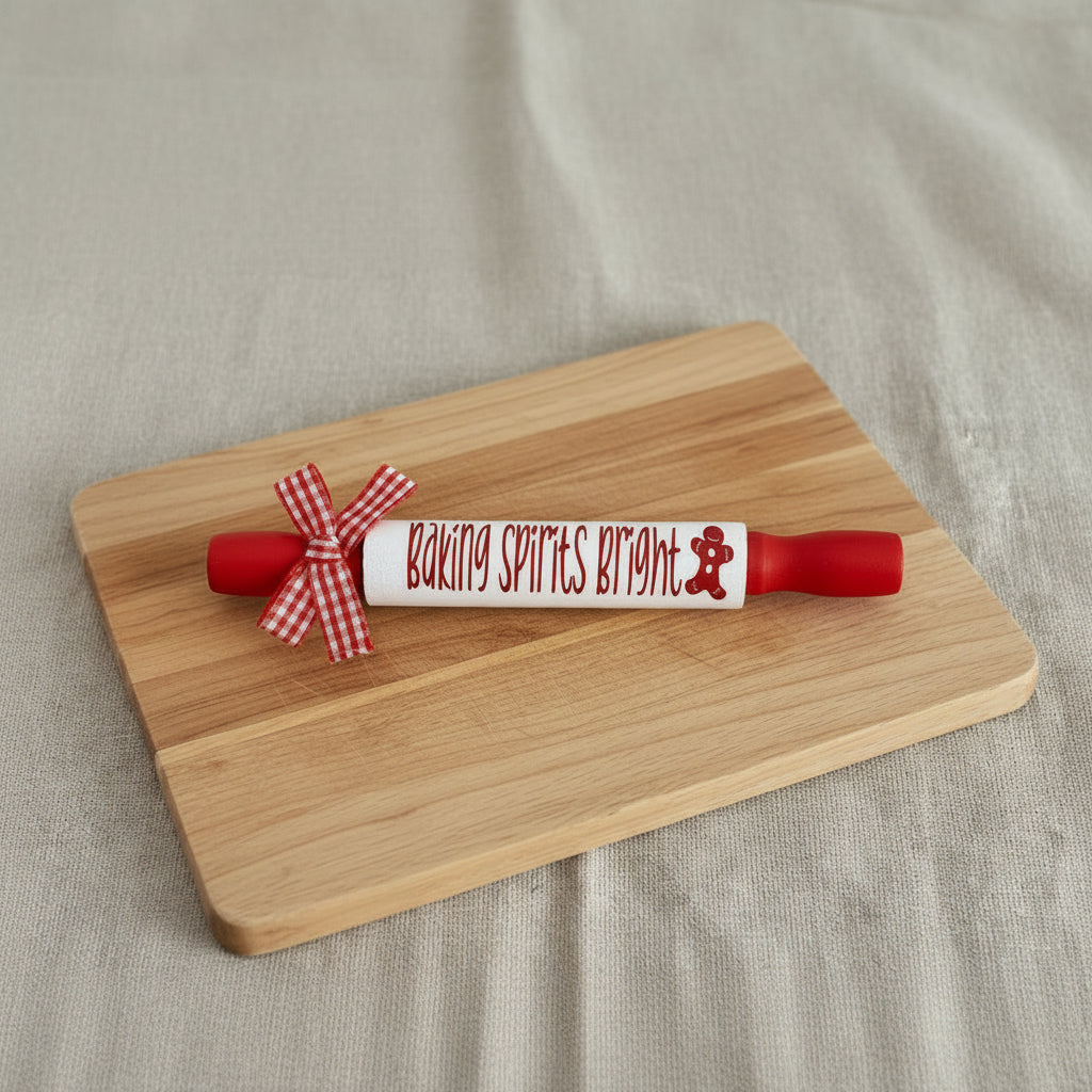 A red rolling pin with a white center section that reads 'Baking Spirits Bright' in red script, decorated with a red and white gingham bow on one end. The rolling pin is placed on a beige linen background with the text 'Sarah's Shop' underneath.