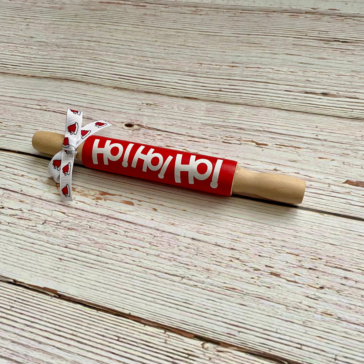 A wooden rolling pin with a red handle printed with 'Ho Ho Ho!' in white, tied with a white ribbon featuring red hearts, placed on a whitewashed wooden surface.