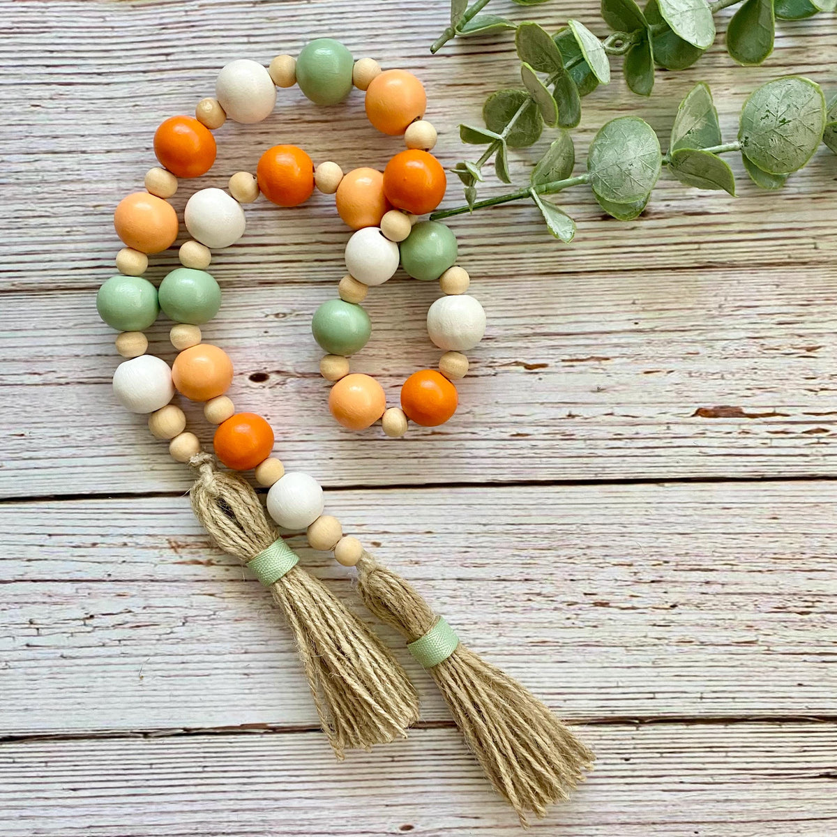 A beaded garland with two tassels, featuring orange, sage green, cream, and natural wood beads, styled on a rustic white-washed wooden surface with eucalyptus leaves.