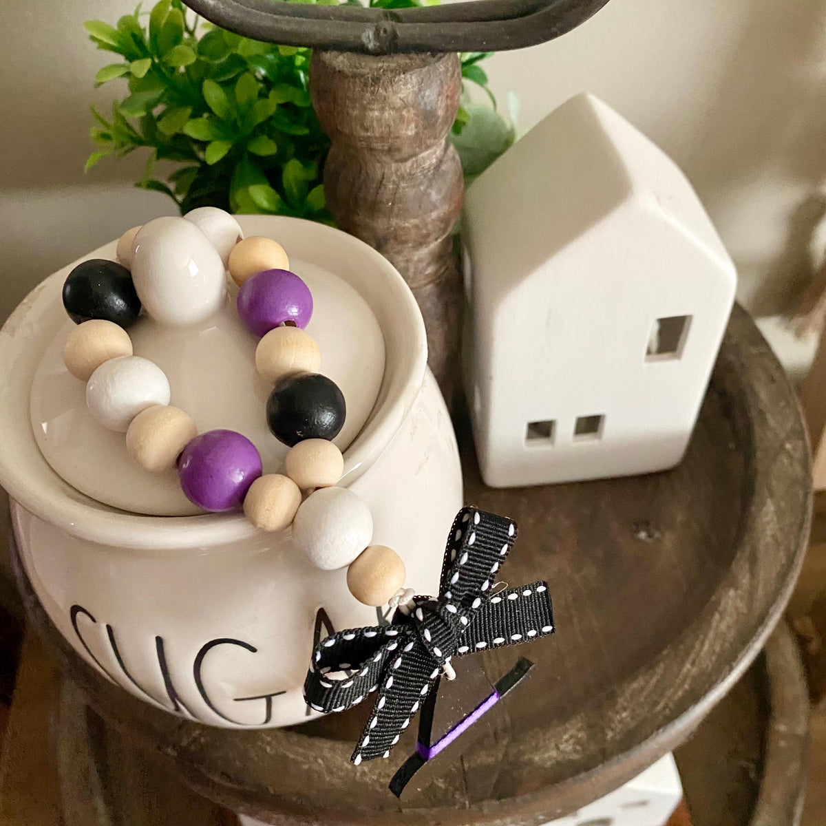 A decorative beaded garland with black, white, beige, and purple wooden beads, styled on a ceramic sugar bowl with a black and white ribbon bow, set on a rustic wooden table.