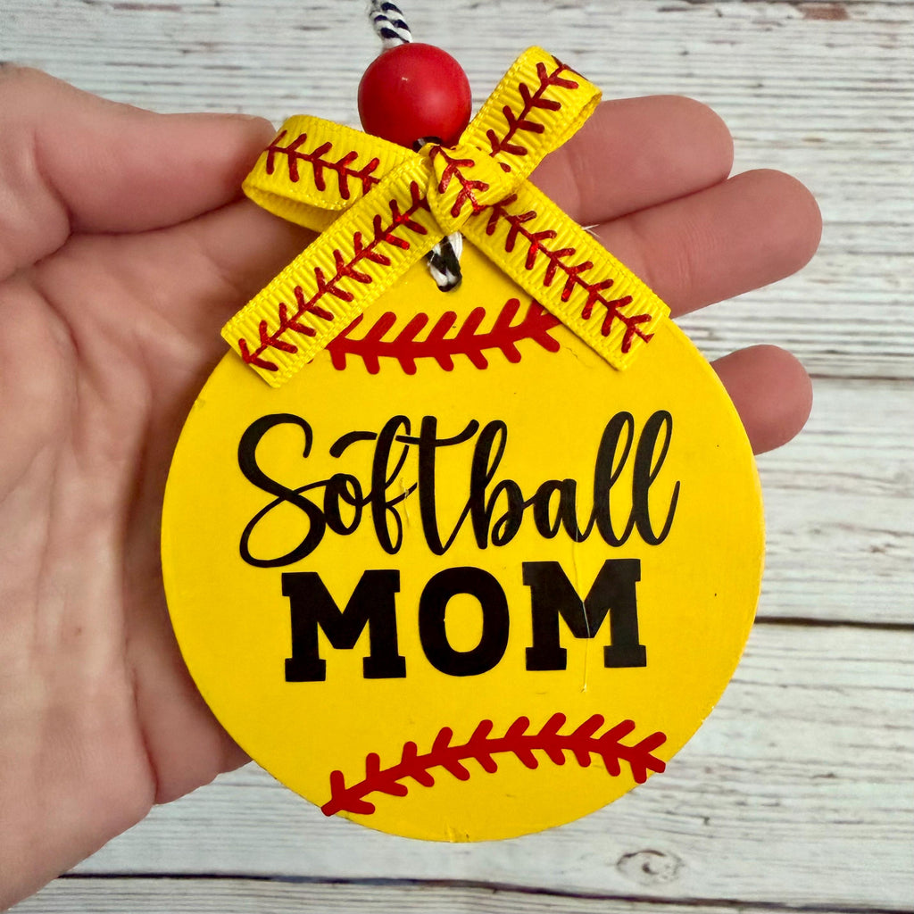 A hand holds a round yellow softball-themed ornament with the words 'Softball MOM' in black lettering and red seam-style details, plus a yellow ribbon bow and red bead at the top, against a whitewashed wood background.