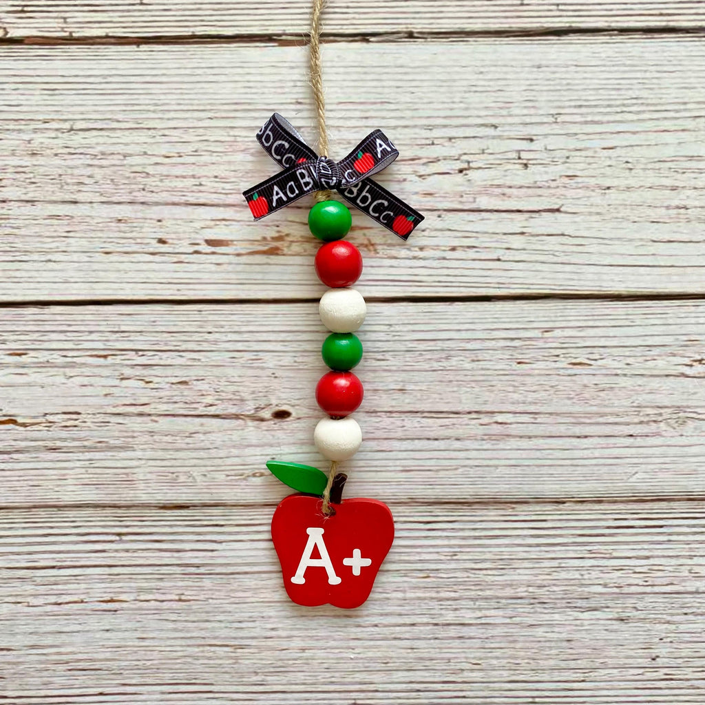 A decorative hanging ornament shaped like an apple with a red body, white leaf, green stem, and white A+ lettering. The ornament is strung with red, green, and white beads, a black ribbon with white text, and hangs from a natural twine string against a whitewashed wood plank background.