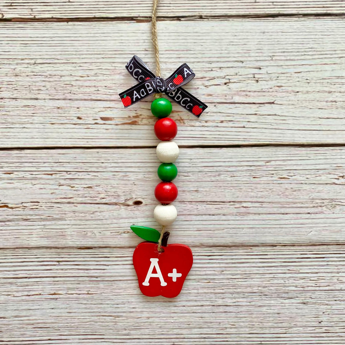 A decorative hanging ornament shaped like an apple with a red body, white leaf, green stem, and white A+ lettering. The ornament is strung with red, green, and white beads, a black ribbon with white text, and hangs from a natural twine string against a whitewashed wood plank background.