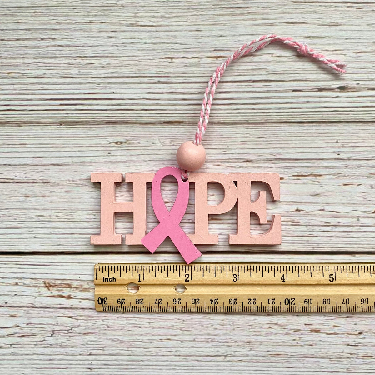 A wooden 'HOPE' word art ornament with a pink ribbon integrated into the letter 'O', hanging from a pink and white twine string with a pink bead. The ornament is displayed on a weathered white wood surface above a wooden ruler to show scale.