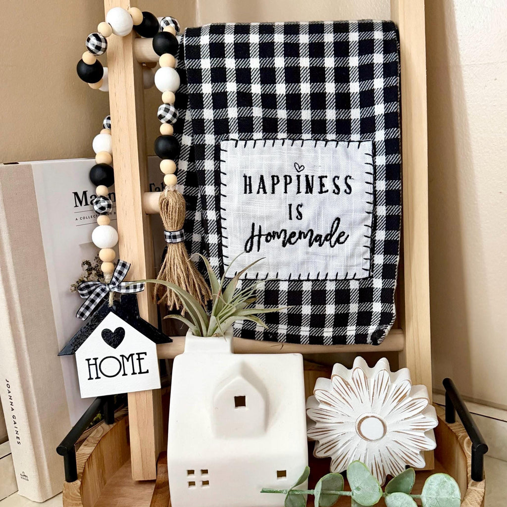 A styled home decor scene featuring a black and white buffalo plaid throw blanket draped over a wooden stand, with a central patch reading “HAPPINESS IS Homemade.” A black-and-white bead garland, a small house-shaped “HOME” sign, a white ceramic house candle holder, and other rustic decor are arranged around it on a tray against a beige wall.