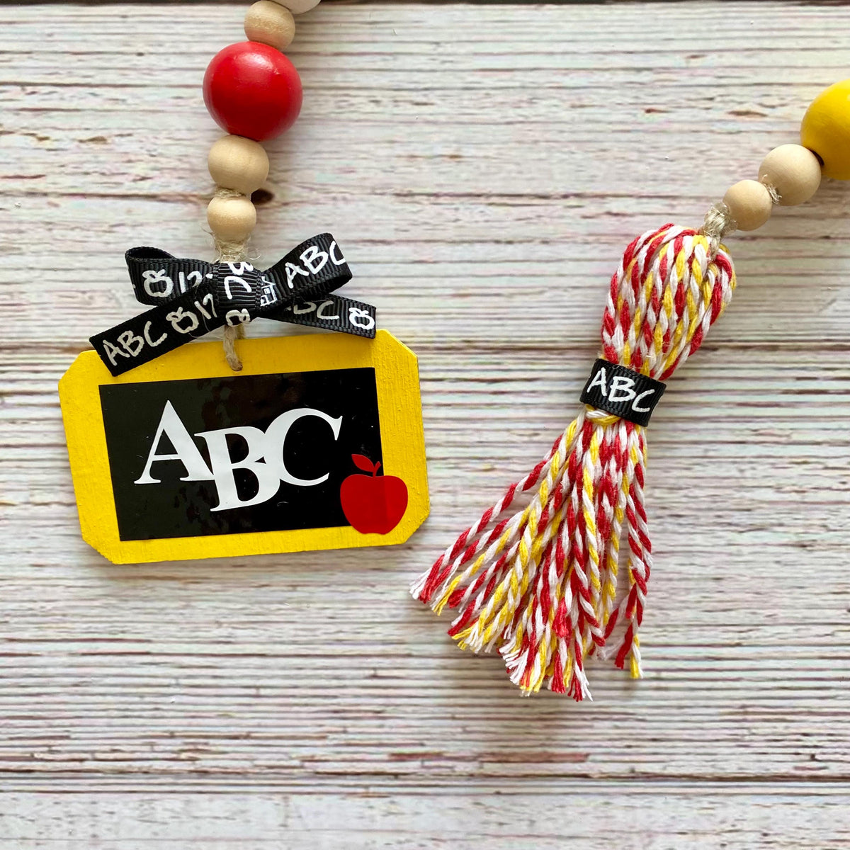 School-themed beaded garland with a yellow chalkboard charm, black and white 'ABC' ribbon, red apple detail, and a red, yellow, and white tassel on a whitewashed wood background.
