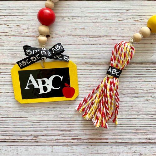 School-themed beaded garland with a yellow chalkboard charm, black and white 'ABC' ribbon, red apple detail, and a red, yellow, and white tassel on a whitewashed wood background.
