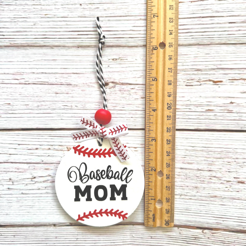 A round white 'Baseball MOM' ornament styled like a baseball with red stitching, a red bead, and a black-and-white string, shown next to a wooden ruler on a whitewashed wood background.