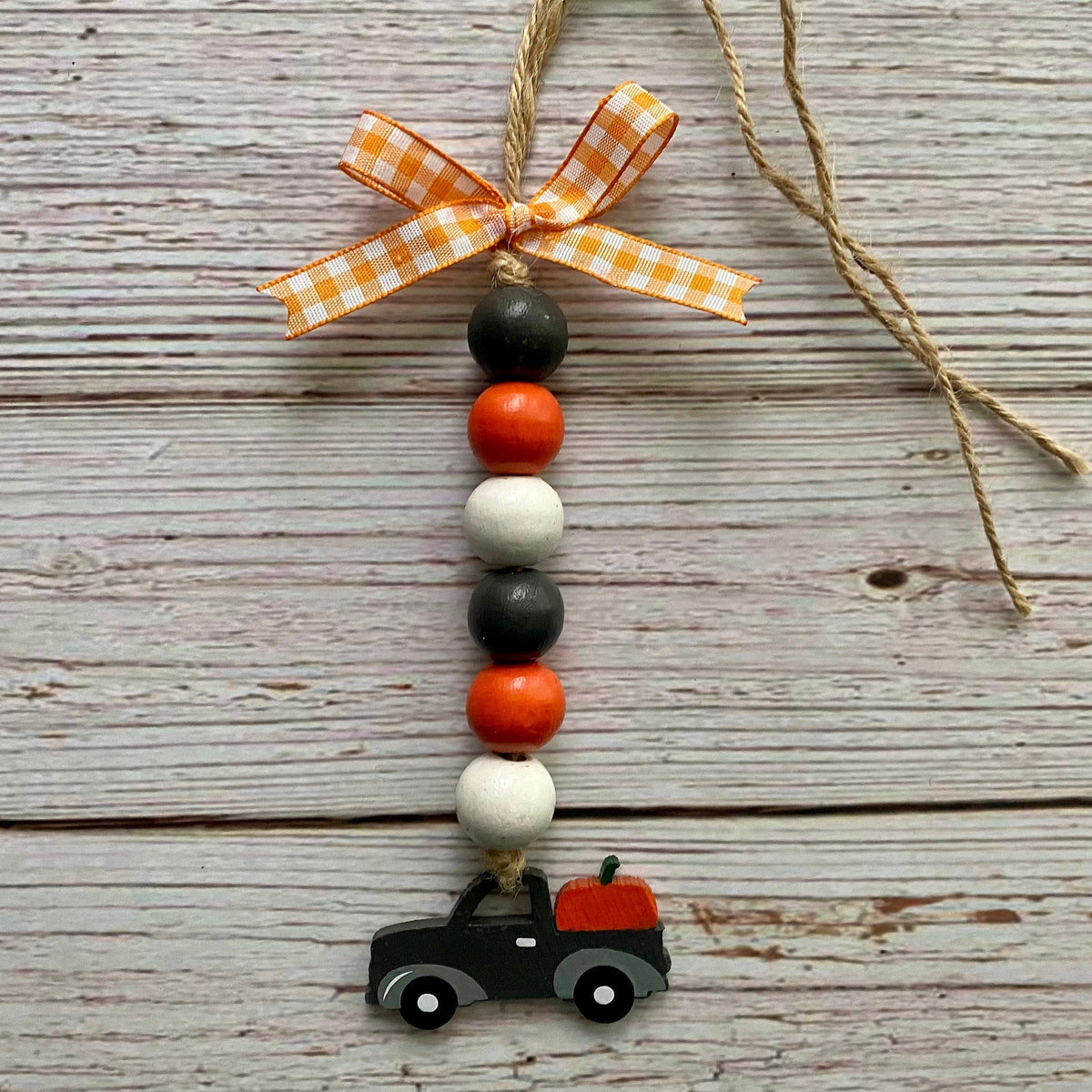 A rustic wooden bead garland ornament featuring orange, black, and white beads, a small painted black truck with an orange bed, and an orange and white gingham ribbon bow at the top, displayed on a weathered gray wood plank background.