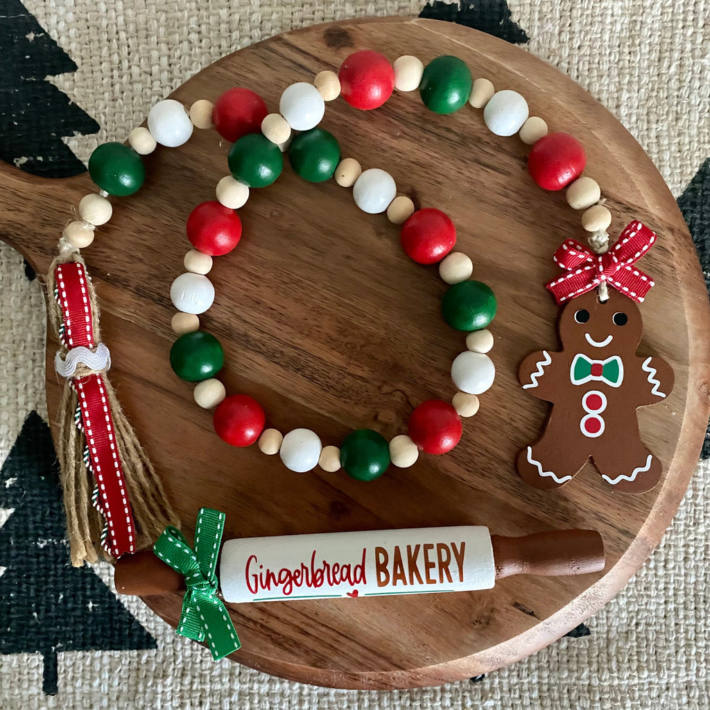A holiday-themed beaded garland set displayed on a round wooden board. The garland features red, green, and white wooden beads, natural wood beads, a red and white ribbon tassel, and a gingerbread man ornament.