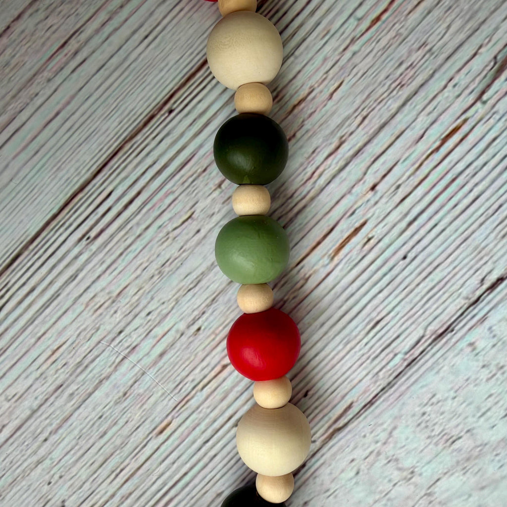 A vertical wooden bead garland with alternating matte natural wood beads, painted green beads, and one prominent red bead, laid on a whitewashed wood plank surface.