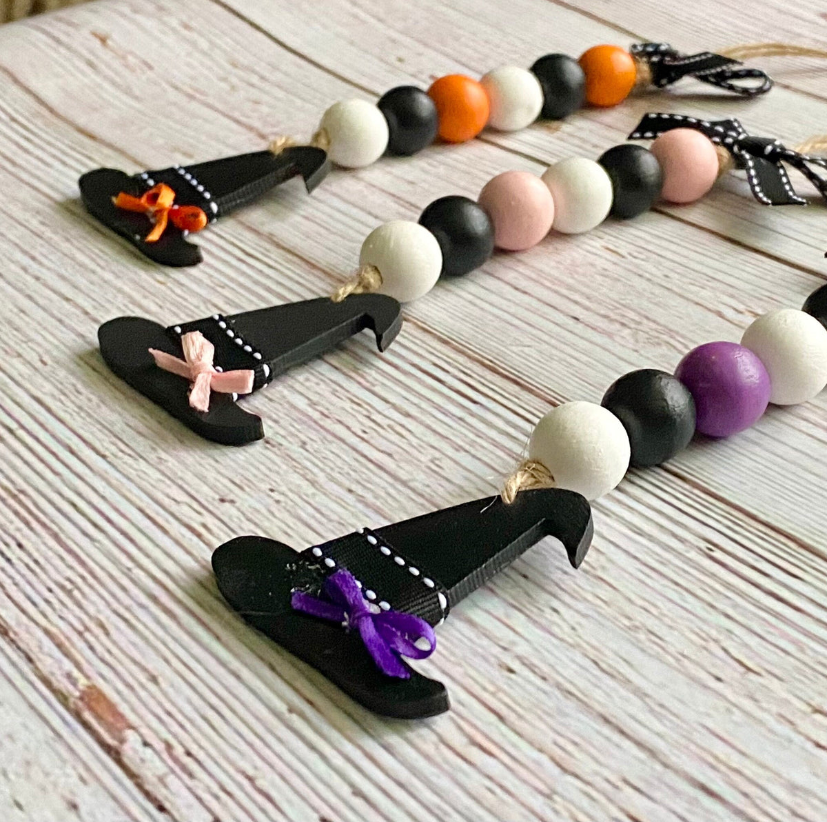 Three wooden witch hat bead garlands with black hats, colorful wooden beads, and ribbon bows, arranged on a whitewashed wooden surface.