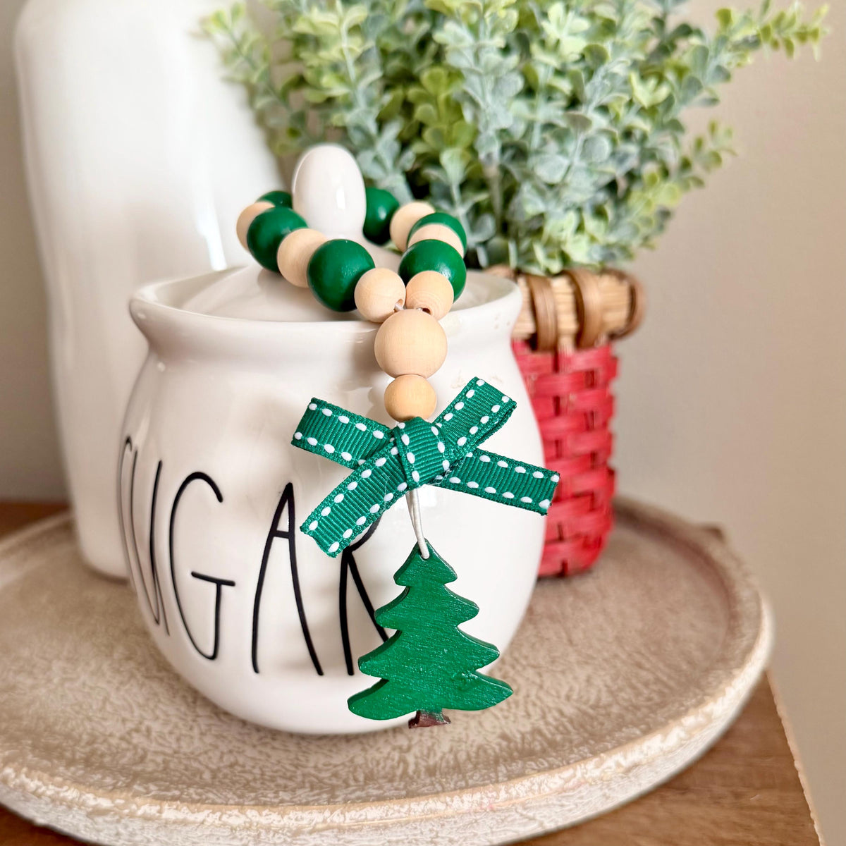 A holiday-themed beaded garland with green and natural wood beads, a green ribbon bow, and a wooden Christmas tree ornament, draped over a white ceramic sugar canister.