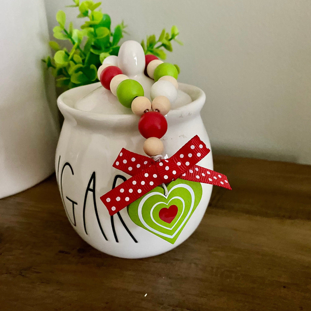 A white ceramic sugar bowl decorated with black lettering, a red and green heart tag, and a red beaded garland, sitting on a wooden table next to another white ceramic container.