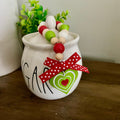 A white ceramic sugar bowl decorated with black lettering, a red and green heart tag, and a red beaded garland, sitting on a wooden table next to another white ceramic container.