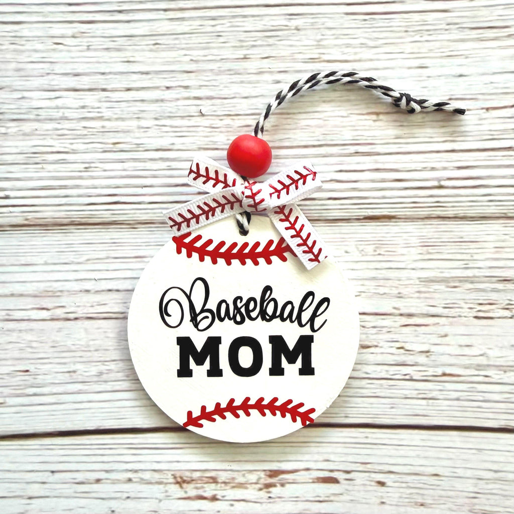 A round white ornament designed like a baseball with red stitching, the words 'Baseball MOM' in black script and block lettering, a red bead, and a black-and-white twine loop, photographed on a whitewashed wood surface.