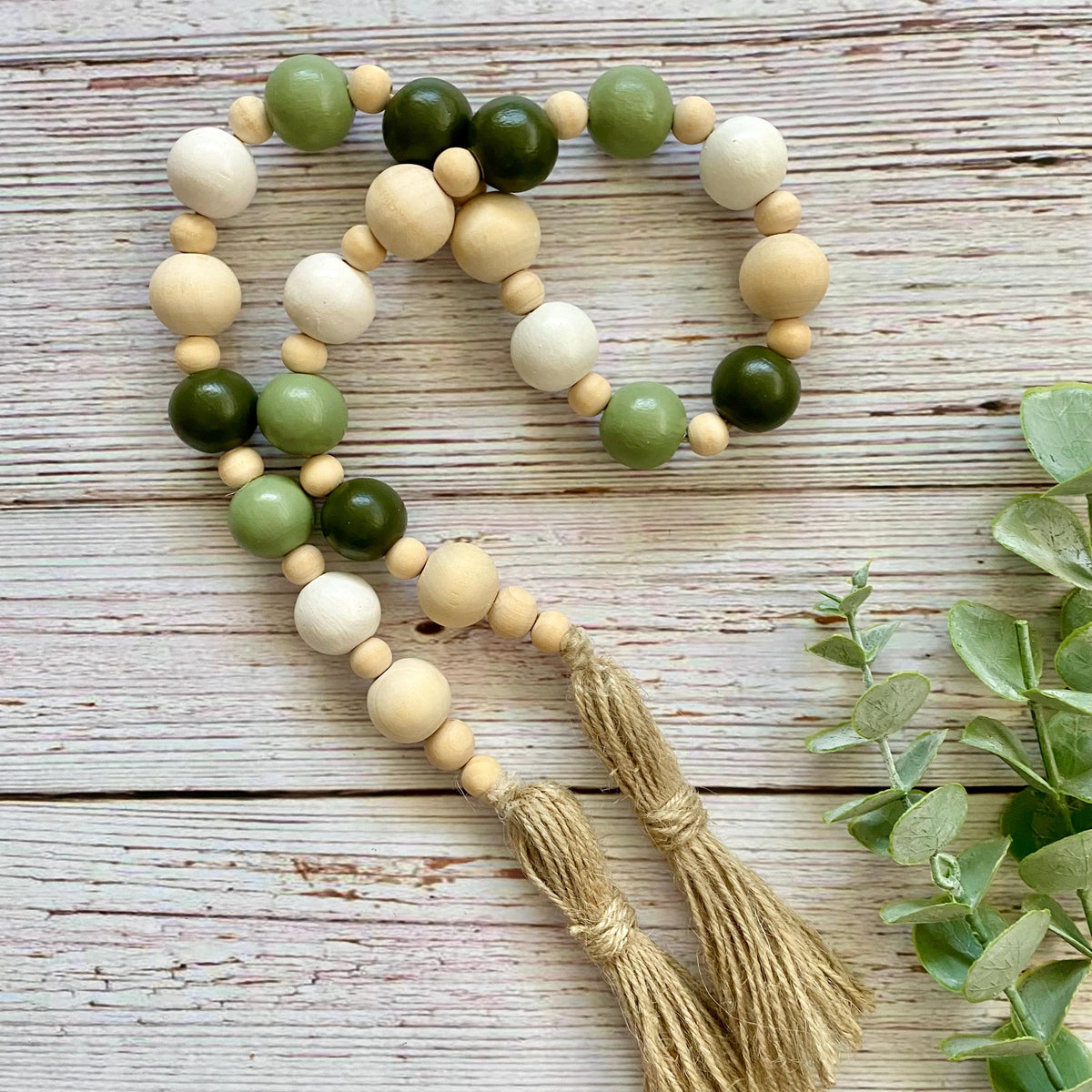 A wooden bead garland with two braided tassels, featuring natural light wood, sage green, olive green, and cream beads, arranged on a rustic whitewashed wooden surface with eucalyptus leaves.