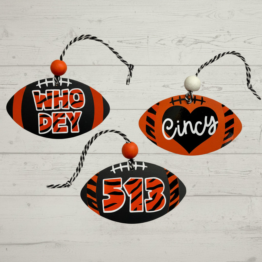 Three football-shaped hanging ornaments on a light wood background. One ornament is black with orange stripes and the word 'WHO DEY' in white and orange.