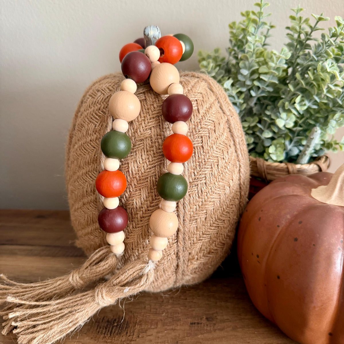 A decorative wooden bead garland with orange, green, burgundy, and natural beige beads, draped over a woven pumpkin on a wooden surface with a potted plant in the background.