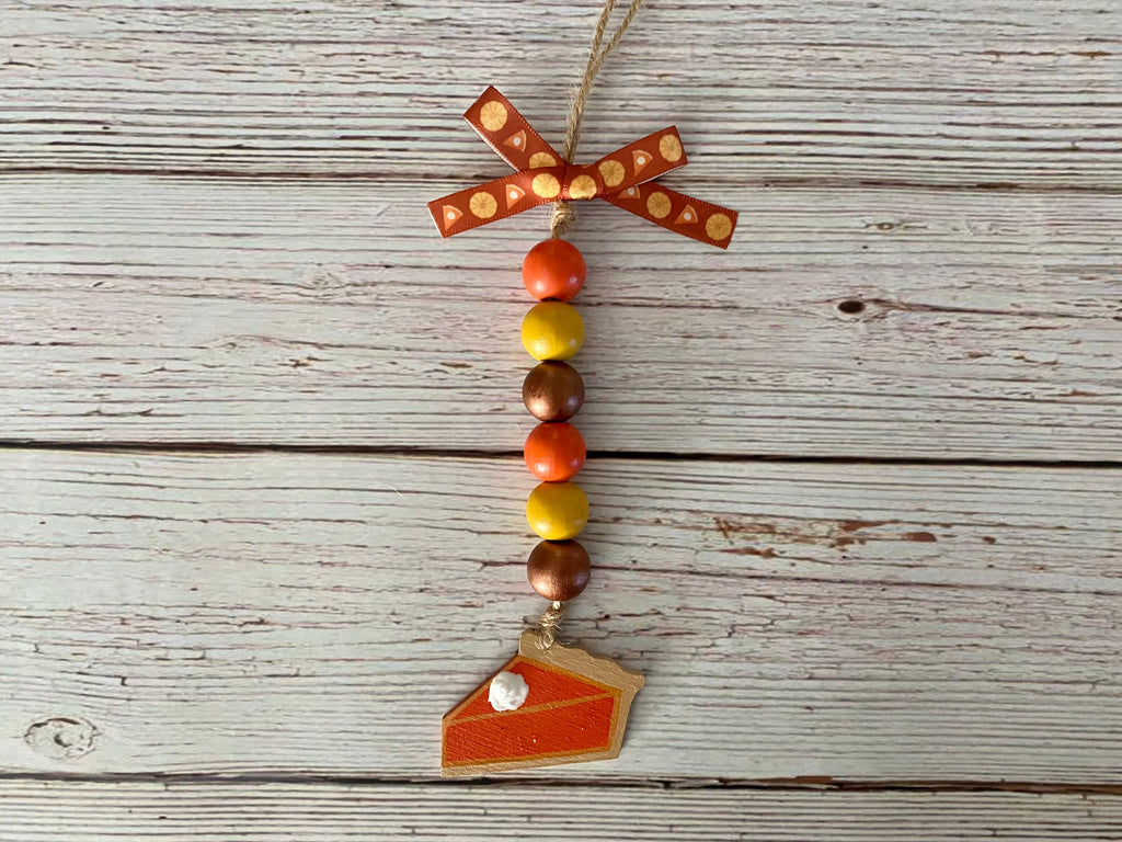 An autumn-themed beaded ornament featuring a patterned orange and brown bow, colorful wooden beads in shades of orange, yellow, and brown, and a small wooden slice of pumpkin pie hanging from a jute string, displayed on a rustic white-washed wooden background.