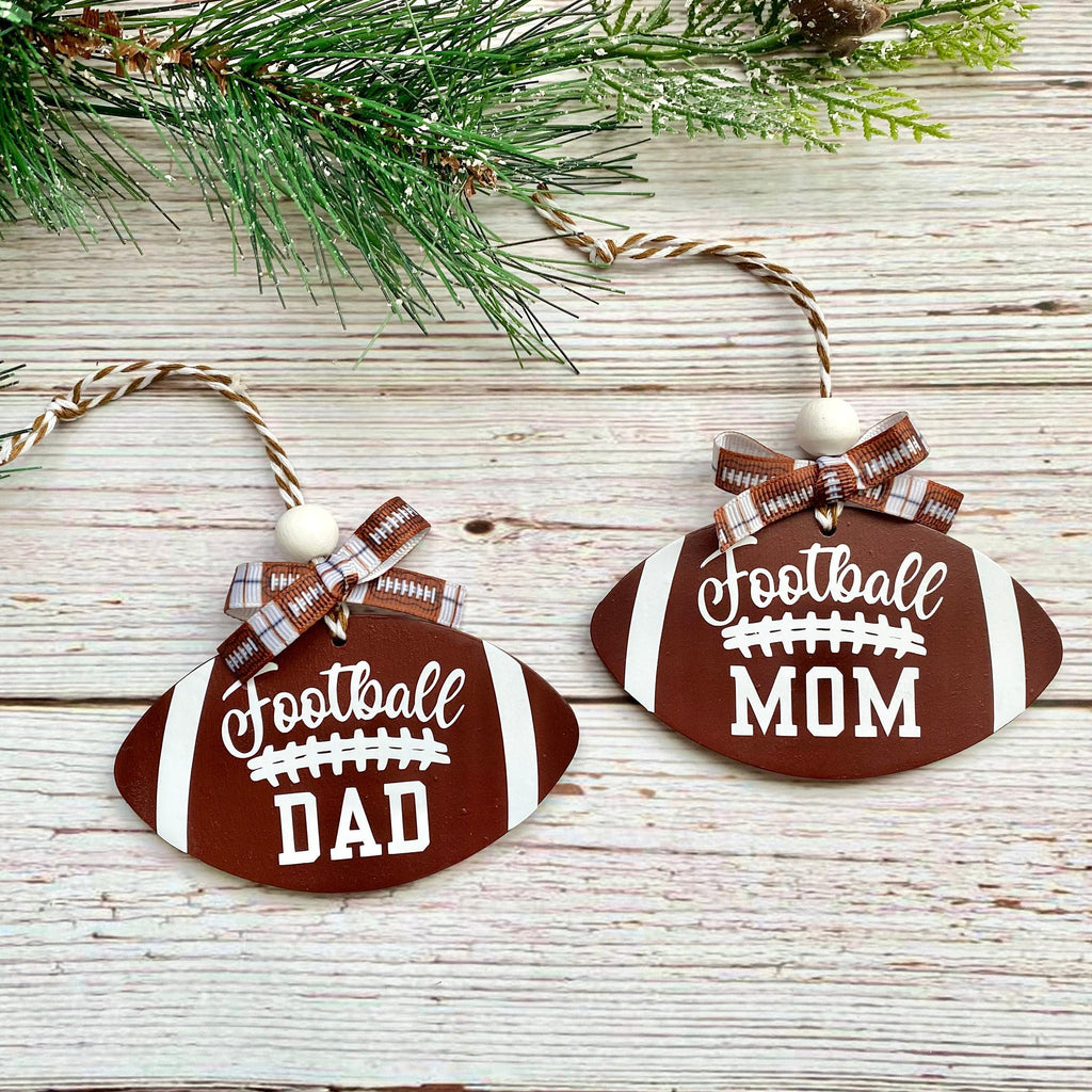 Two football-shaped wooden ornaments on a whitewashed wood surface with pine branches at the top. One ornament reads "Football DAD" and the other reads "Football MOM" in white lettering, both with brown and white plaid bows.