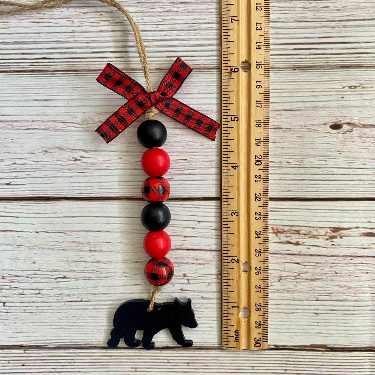 A rustic beaded hanging ornament with a red and black buffalo plaid ribbon bow, alternating red and black beads, and a black bear silhouette at the bottom, shown next to a wooden ruler on a whitewashed wood plank background.