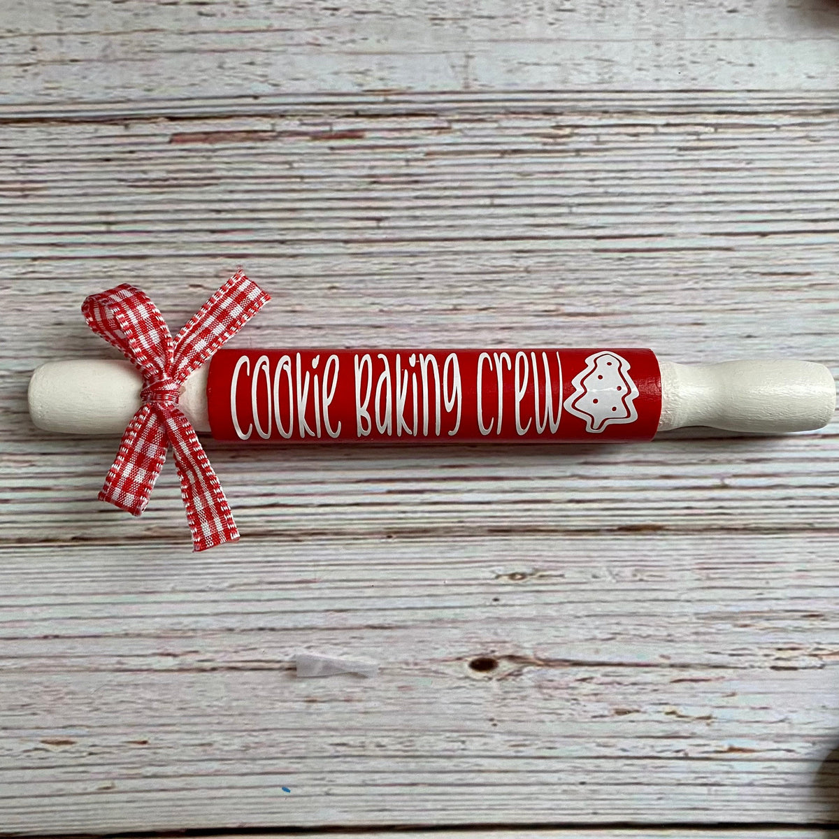 A wooden rolling pin with a red painted center and white lettering that reads 'Cookie Baking Crew' with a white gingerbread cookie outline, tied with a red-and-white gingham ribbon. It is photographed on a whitewashed wood surface with a decorative garland partially visible along the right edge.
