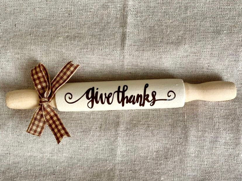 A wooden rolling pin with the words 'give thanks' written in black script on its white-painted handle, tied with a brown and beige gingham ribbon, set against a textured beige linen background.
