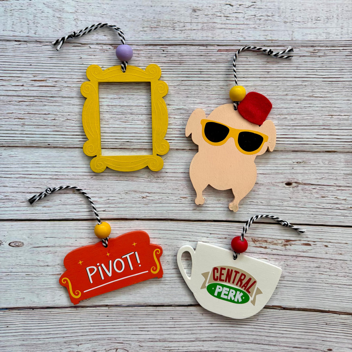 Four novelty Christmas ornaments shaped like the Friends coffee table decor, displayed on a light gray wood background. The ornaments include a yellow picture frame, a turkey wearing yellow sunglasses and a red hat, a orange couch with PIVOT!, and a white Central Perk coffee cup.