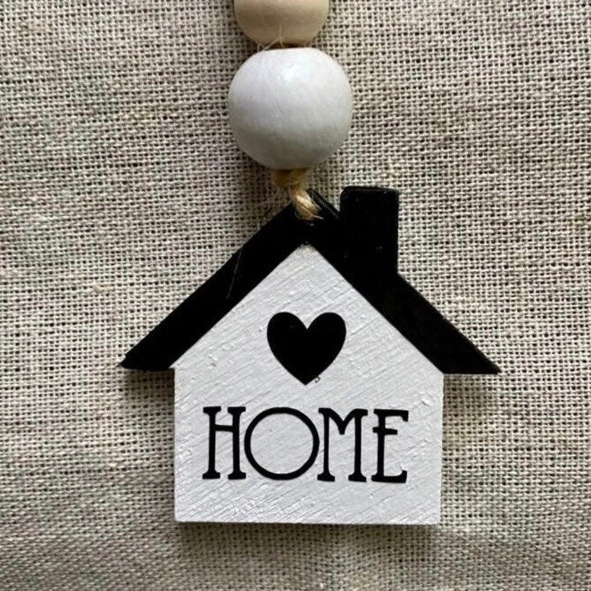 A small house-shaped hanging ornament with a black roof, black heart, and the word HOME in black lettering on a white body, suspended from wooden beads against a beige linen background.