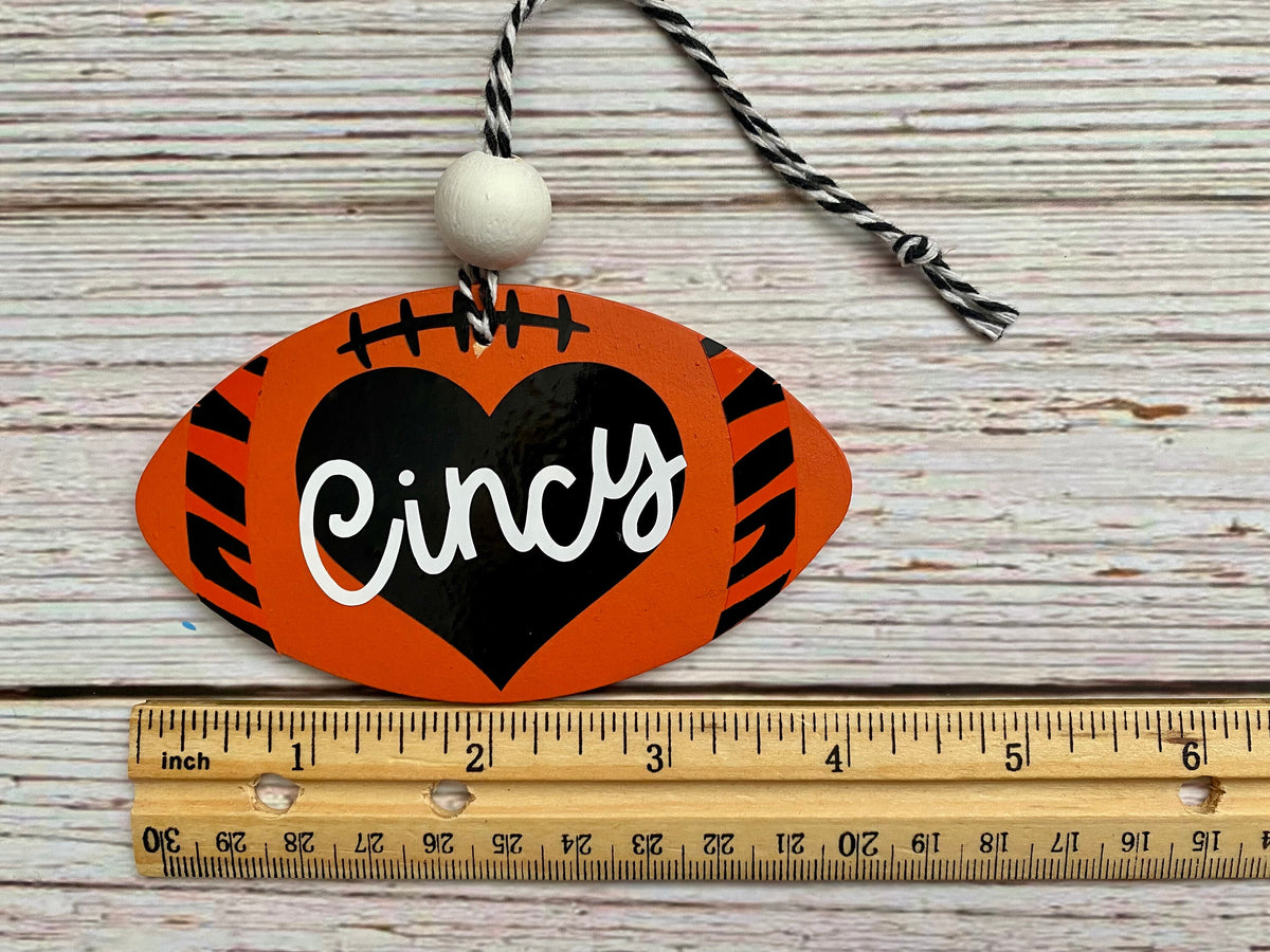 A football-shaped wooden ornament with orange tiger-stripe sides, a black center, white script text reading 'Cincy,' and a black-and-white hanging cord. A wooden ruler is placed below it on a whitewashed wood surface to show size.
