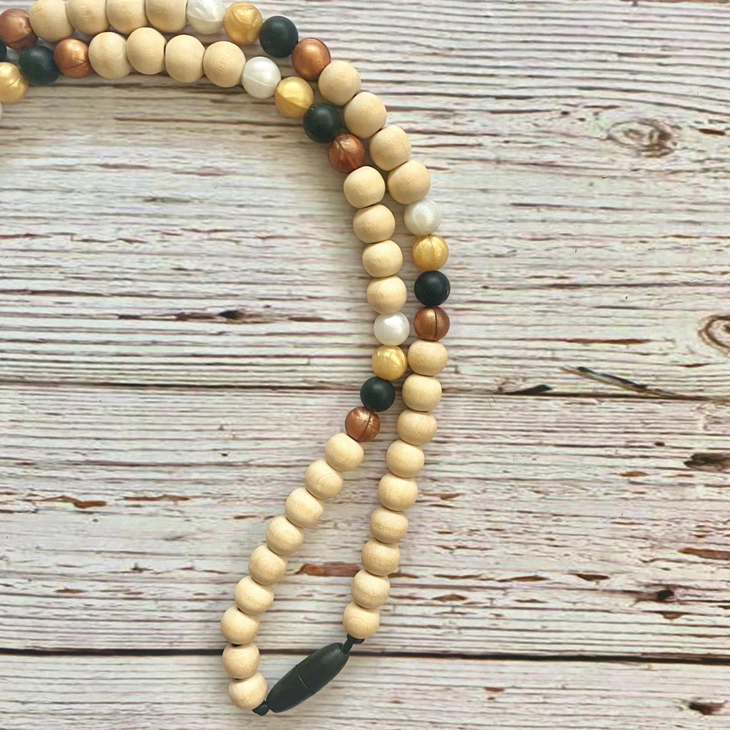 A beaded necklace made of natural wood beads in light tan, medium brown, and dark brown, accented with a few dark green and white beads, laid out on a rustic white-washed wooden surface.