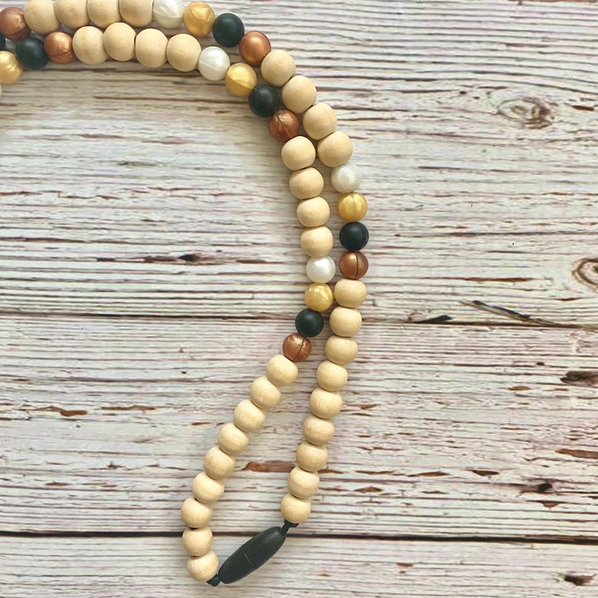 A beaded necklace made of natural wood beads in light tan, medium brown, and dark brown, accented with a few dark green and white beads, laid out on a rustic white-washed wooden surface.