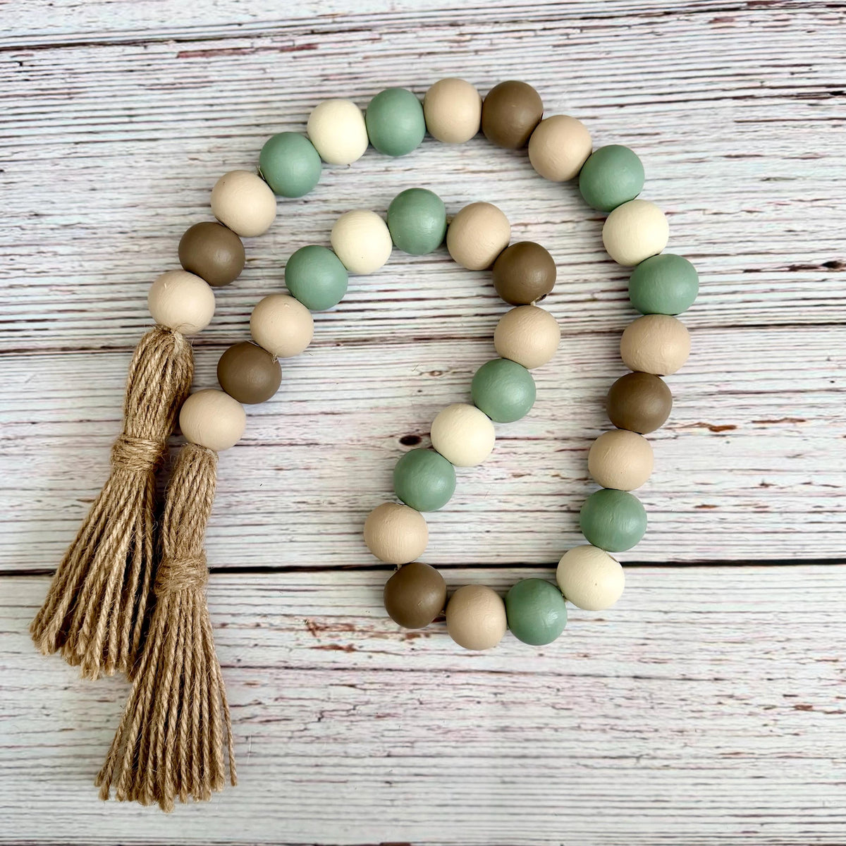 A beaded garland made of matte wooden beads in sage green, cream, tan, and medium brown, with two knotted jute tassels, arranged on a whitewashed wood surface.