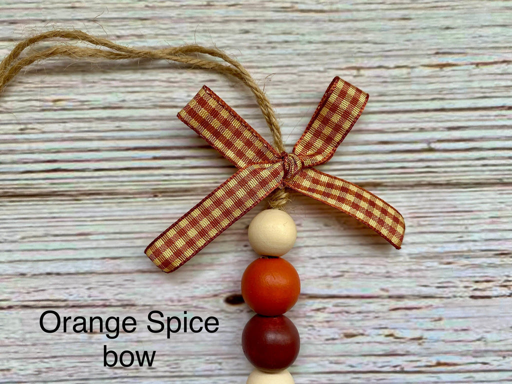 A decorative hanging ornament or bow with a red-and-cream gingham ribbon bow, three hanging wooden beads in cream, orange, and dark red, and twine for hanging, shown on a whitewashed wood background with the text 'Orange Spice bow'.