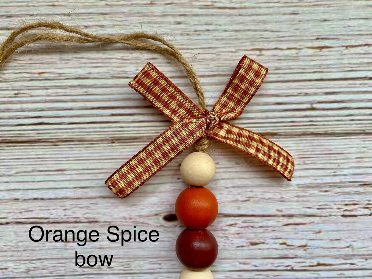 A decorative hanging ornament or bow with a red-and-cream gingham ribbon bow, three hanging wooden beads in cream, orange, and dark red, and twine for hanging, shown on a whitewashed wood background with the text 'Orange Spice bow'.