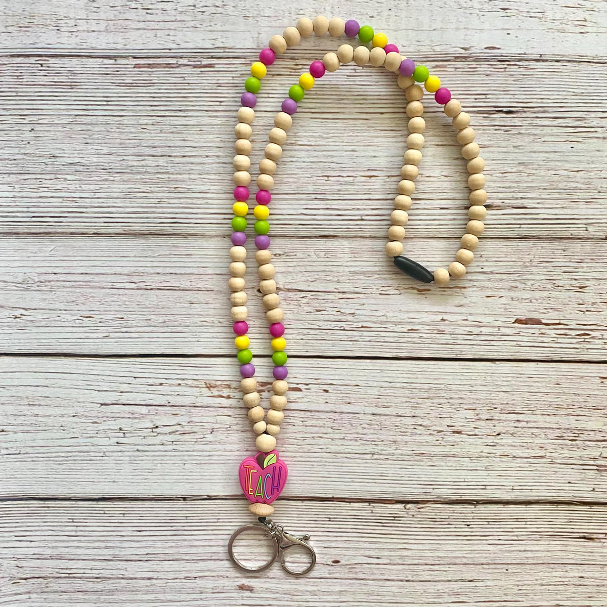 A beaded keychain or lanyard made of natural wood beads with colorful accents in pink, green, yellow, and purple, featuring a pink heart charm with colorful stripes and a silver metal clasp, displayed on a rustic white-washed wooden background.
